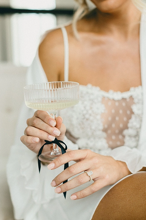 Bride holding champagne in a coupe glass tied with a black ribbon, showing engagement ring and nude manicure in soft window light