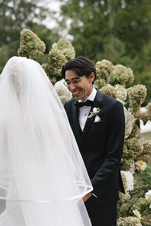 Wedding vows as bride in white veil and groom in black tuxedo with glasses hold hands under a floral arch in a garden setting