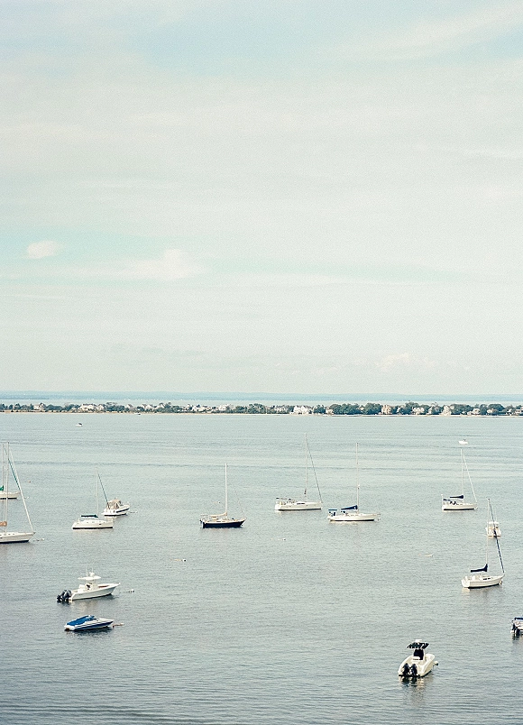 Harbor scenery with sailboats on water and a few motorboats anchored in a calm bay, distant coastal houses beneath a cloudy sky
