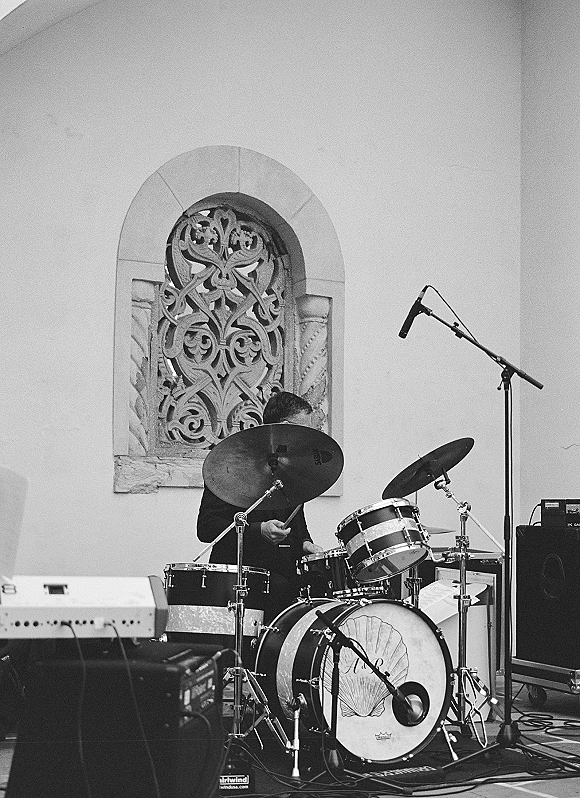 Wedding band drummer at a wedding band setup with drum kit and cymbals, near a microphone stand by an arched window indoors