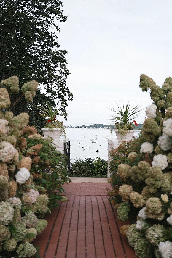 Ceremony aisle lined with white hydrangea aisle decor in large planters along a brick walkway, leading to a gate with sailboats beyond