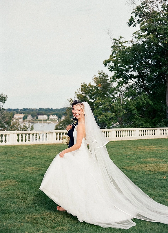 Couple portrait of bride in strapless ball gown and cathedral veil with groom in tuxedo, holding bouquet by stone balustrade near water