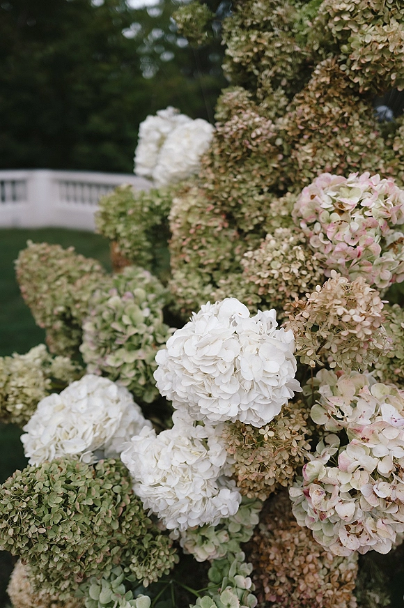 Wedding floral arrangement with hydrangea wedding flowers in white, blush, and green set on a lawn beside garden greenery and a white fence