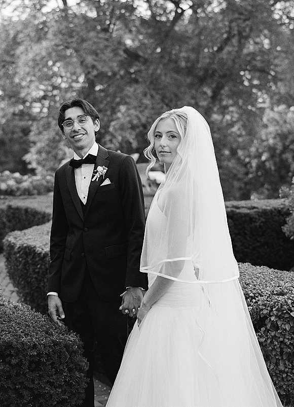 Couple portrait in a black and white wedding portrait style, bride in veil and strapless gown holding hands with tuxedoed groom in glasses in a hedge garden