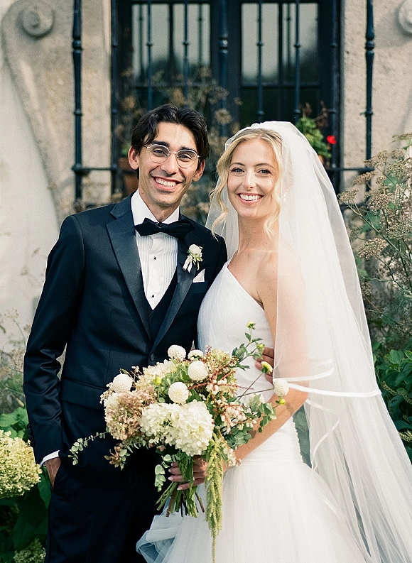 Couple portrait of bride and groom smiling, holding a white and green bouquet, her veil draped over a strapless dress in a lush garden by an iron-barred window