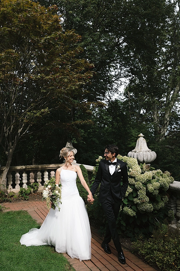 Couple portrait of bride and groom holding hands, she carries a bouquet, walking by a stone balustrade in a garden setting.