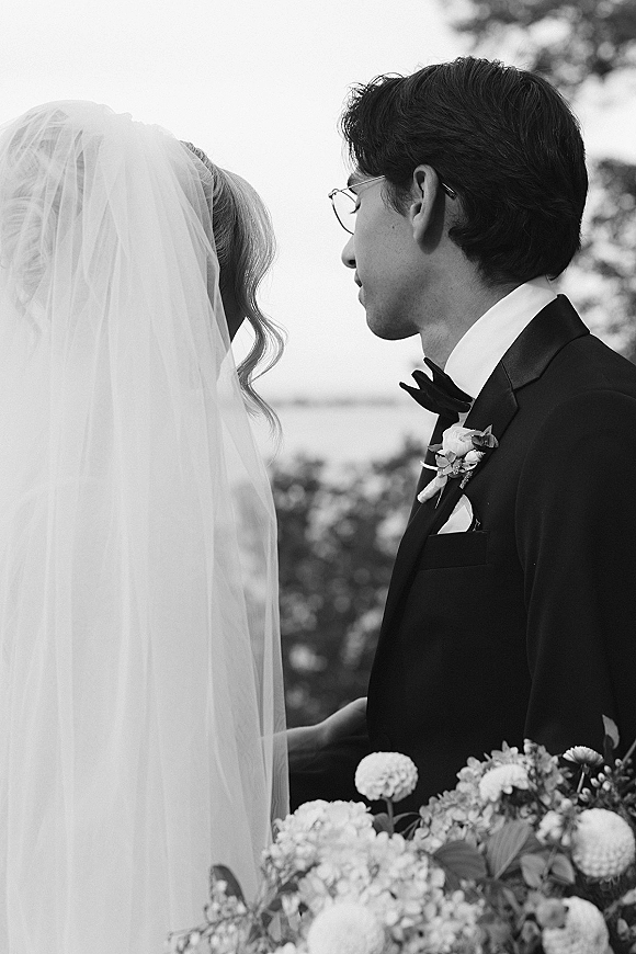Wedding couple portrait in black and white, bride in long veil beside groom in tuxedo and glasses, trees and distant water behind