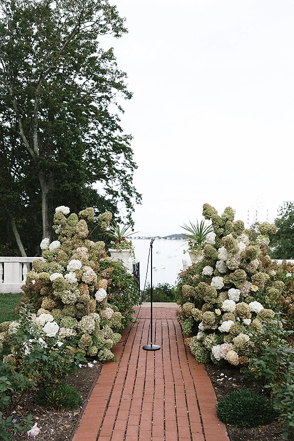 Ceremony aisle decor with hydrangea ceremony flowers and greenery lining a brick walkway, with a microphone stand by a waterfront terrace view
