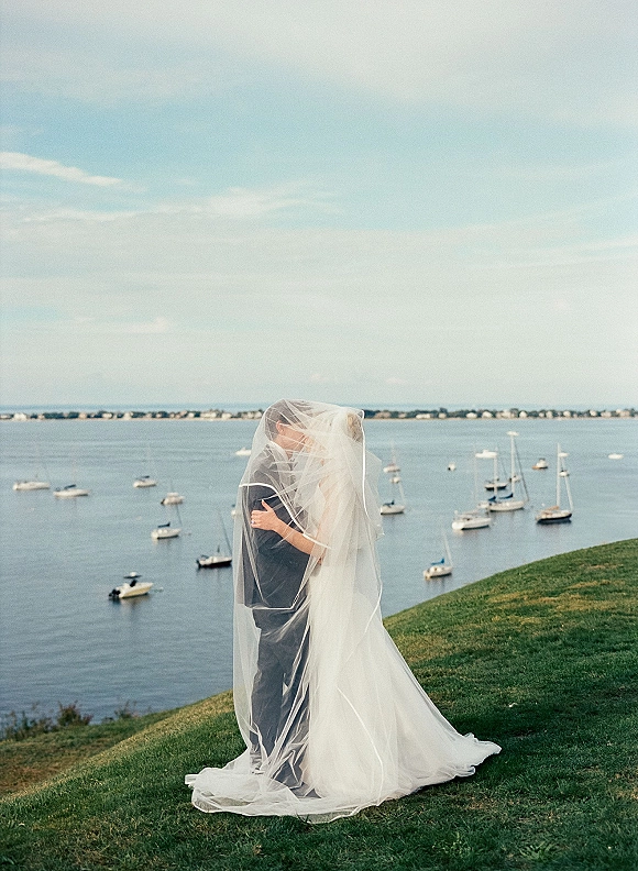 Wedding kiss portrait of bride and groom under veil, veil kiss photo with windswept veil on a grassy hill above ocean and sailboats
