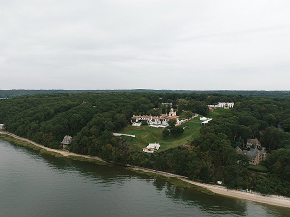 Wedding venue aerial showing an estate wedding venue with a white tent on a grassy lawn beside a lake shoreline and forest trees