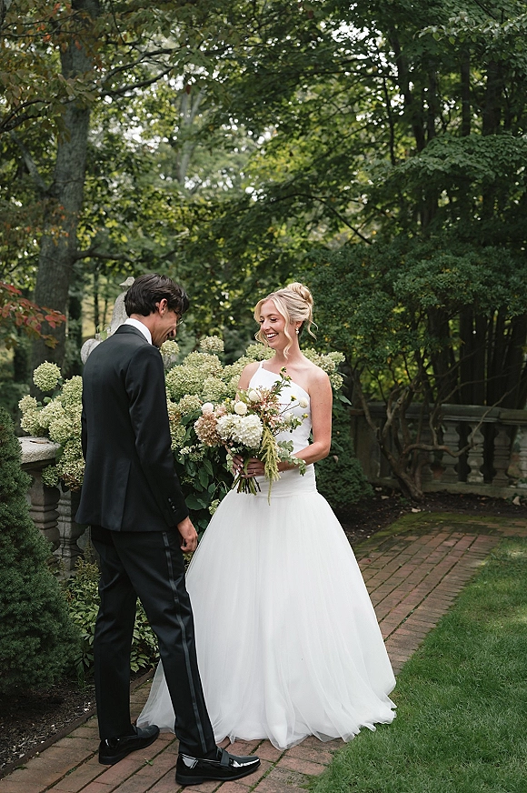 First look moment in a garden as bride in a strapless gown holds a bouquet while laughing with groom in a tux beside hydrangeas
