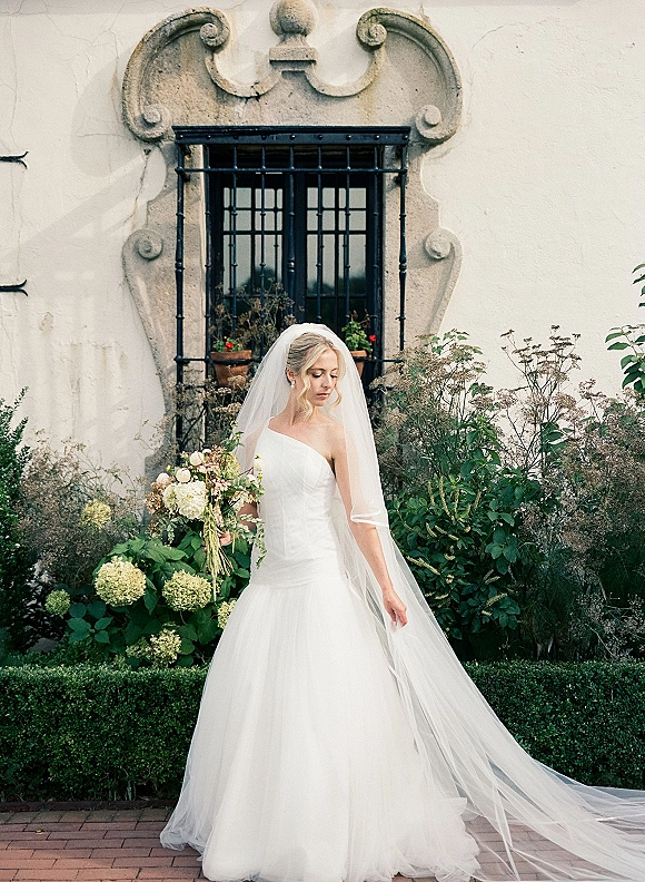 Bridal portrait of a bride in a strapless gown with a cathedral veil, holding a bouquet by a stucco wall and ornate window frame