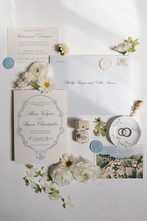 Wedding invitation suite with letterpress wedding invitations, blue wax seals, rings, and white flowers on a textured surface with shadows