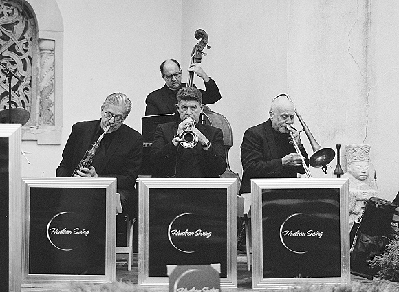 Wedding band performing live wedding music on a stage with music stands, saxophone, trumpet, trombone, and upright bass against a white wall