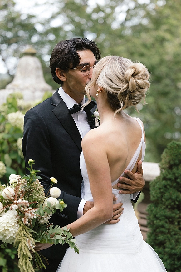 Wedding kiss as bride and groom kissing in a garden by a stone fountain, her one-shoulder dress and white-green bouquet prominent