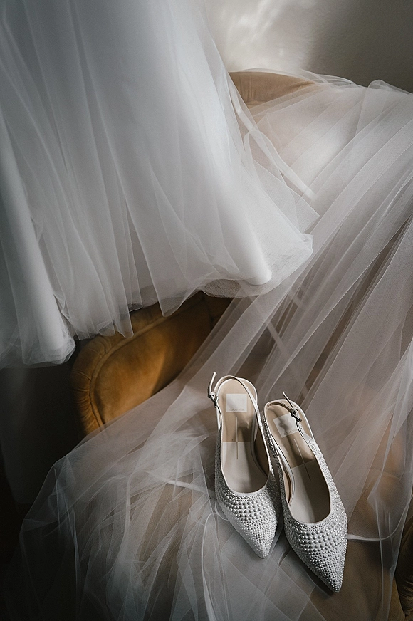 Wedding shoes, pearl wedding heels with pointed toes and ankle straps, resting on an upholstered chair in soft window light