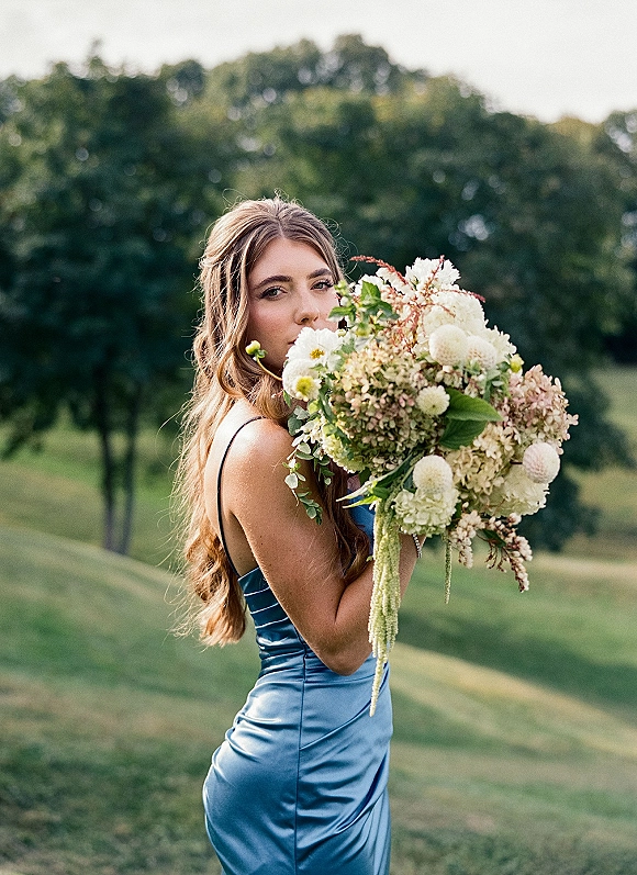 Bridesmaid portrait with an oversized bouquet of mixed cream florals and greenery, posing in a blue satin dress in a grassy field with trees