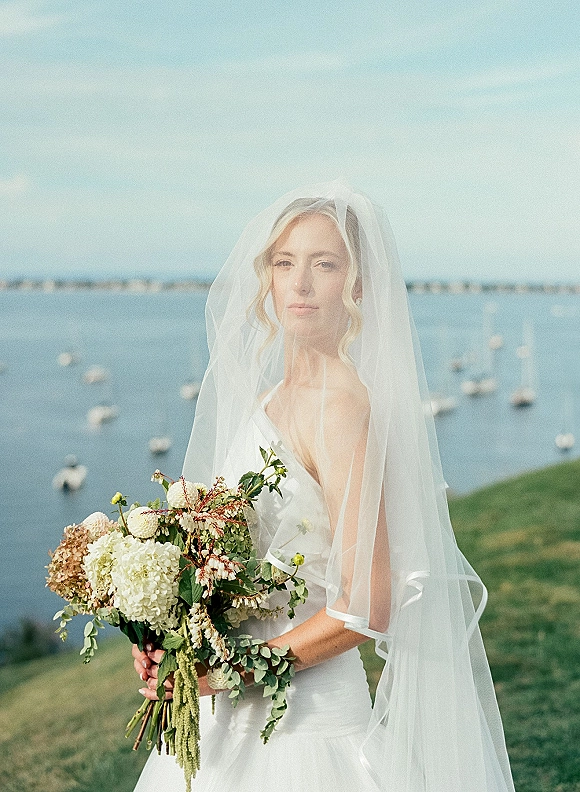 Bridal portrait of a bride holding bouquet with hydrangea and greenery, wearing a long veil and strapless dress by the ocean shore