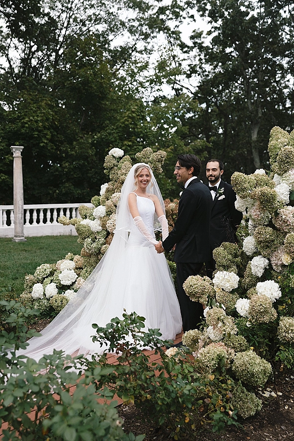 Ceremony moment at an outdoor wedding ceremony with bride and groom holding hands under a white hydrangea arch on a garden lawn