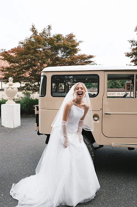 Bridal portrait of a laughing bride in a cathedral veil and off-the-shoulder gown with sheer gloves, beside a vintage SUV outdoors