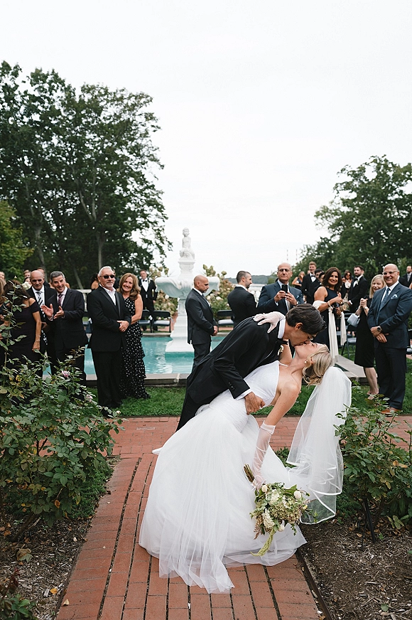 Wedding kiss portrait of groom dipping bride in a dip kiss wedding photo, her veil flowing as guests watch near a garden fountain walkway