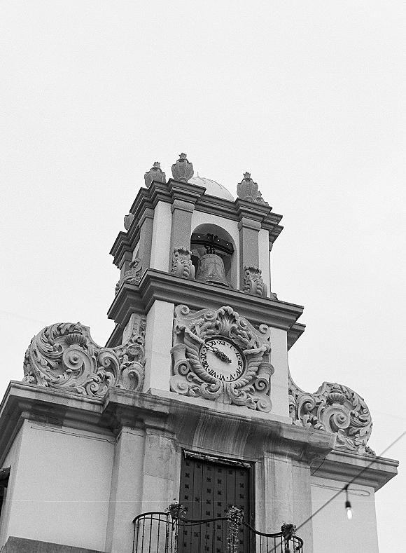Church facade with church clock tower featuring ornate stone carvings and an arched bell opening beneath string lights and sky