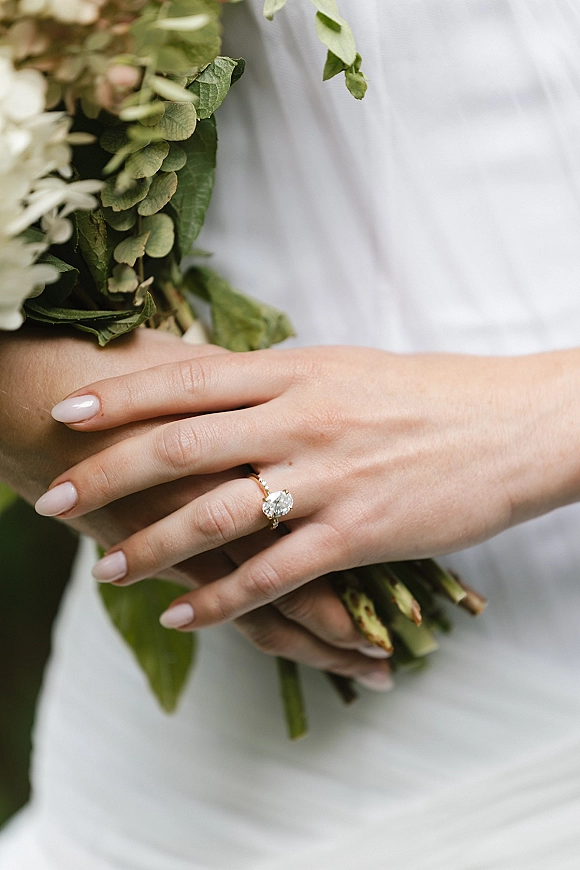 Engagement ring with an oval diamond on a thin gold band, shown on a manicured hand holding a white bouquet with greenery outdoors