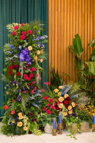 Wedding floral installation ceremony floral backdrop with tropical leaves and roses, pillar candles and baskets against a wood slat wall