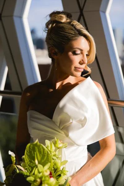 Bridal portrait of a bride in a one-shoulder wedding dress holding a greenery bouquet by modern windows with soft city skyline light