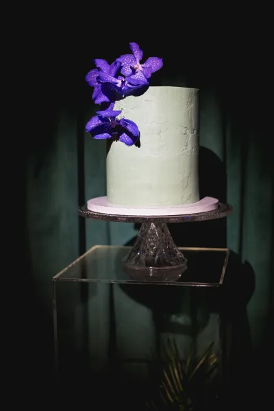 Wedding cake with textured buttercream, single tier design topped with purple orchids on an acrylic pedestal against a dark drape backdrop