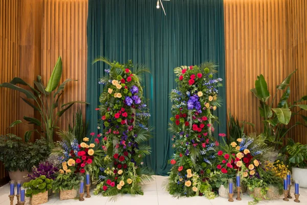 Wedding backdrop decor with teal drapery and colorful floral pillars, tropical greenery and candles set against a wood slat wall indoors