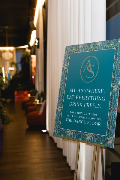 Wedding welcome sign with gold lettering and monogram on an easel, framed by a floral border in a warmly lit indoor hallway