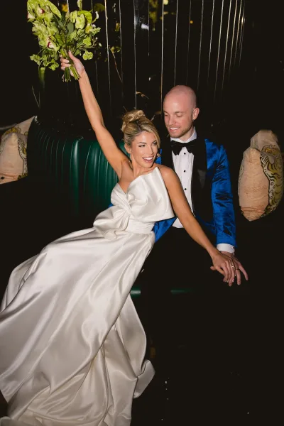 Couple portrait of newlyweds seated as bride holds bouquet up beside groom in blue velvet jacket on a green tufted banquette