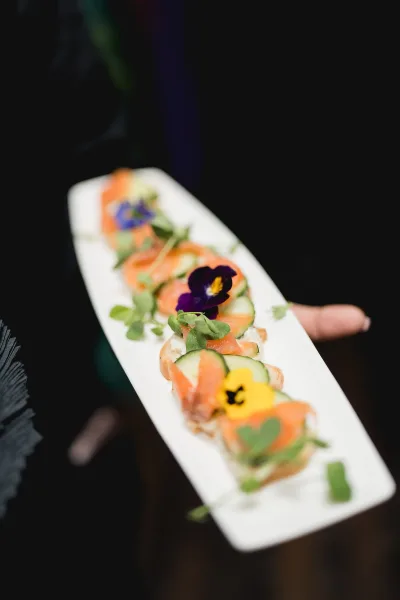 Wedding appetizer crostini with smoked salmon, cucumber, microgreens and edible flower accents on a white platter held by a guest in a blurred crowd
