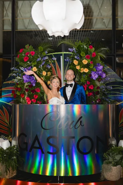 Couple portrait of bride and groom posing under a colorful floral arch, strapless dress and blue velvet tuxedo in a glass-walled venue