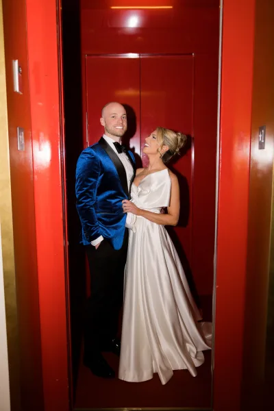 Couple portrait in a red elevator, bride in one-shoulder wedding dress with bow detail gazing at groom in blue velvet tuxedo jacket