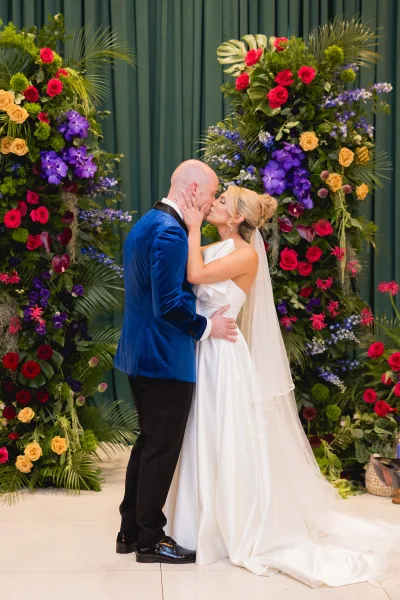 Wedding kiss portrait of bride and groom kissing, her veil and satin dress against a teal wall with tropical florals and greenery backdrop