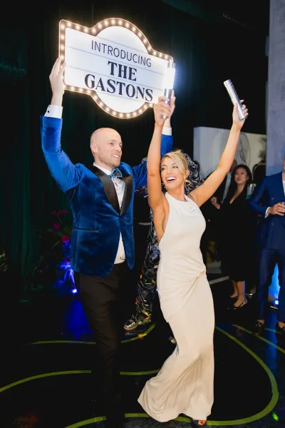 Reception entrance with newlyweds in bride dress and groom blue velvet jacket raising arms by a lighted marquee sign, confetti popping, black drape backdrop