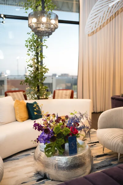 Wedding lounge decor with a white sofa and throw pillows, metallic coffee table floral centerpiece, under a pendant chandelier by tall windows
