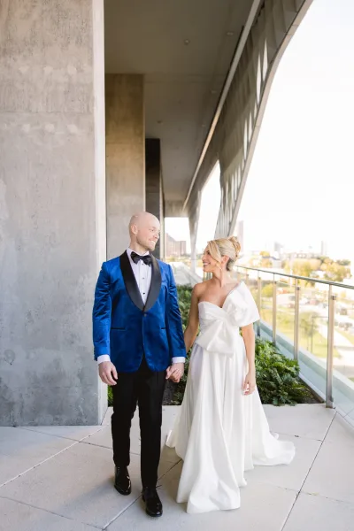 Couple portrait of bride and groom holding hands, her one-shoulder dress and his blue velvet tuxedo on a modern balcony walkway with skyline
