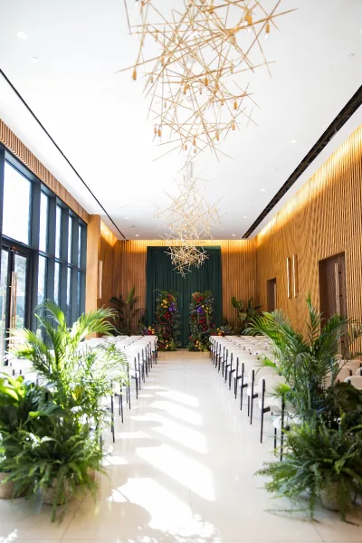 Ceremony setup for an indoor wedding ceremony with white chairs and greenery aisle decor, floral altar and sculptural chandeliers by tall windows