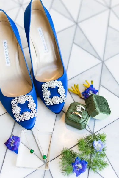 Bridal shoes in blue wedding shoes flat lay with crystal buckles, rings in green velvet box and emerald earrings on white geometric tile floor