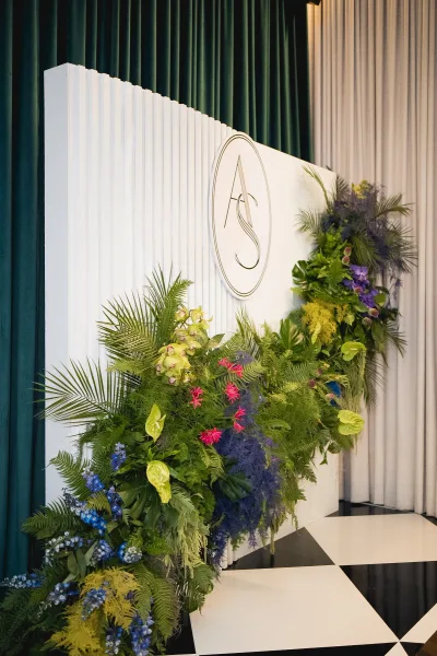 Wedding photo backdrop with a round wedding monogram sign framed by tropical greenery, orchids, and blue-purple flowers on a checkered floor