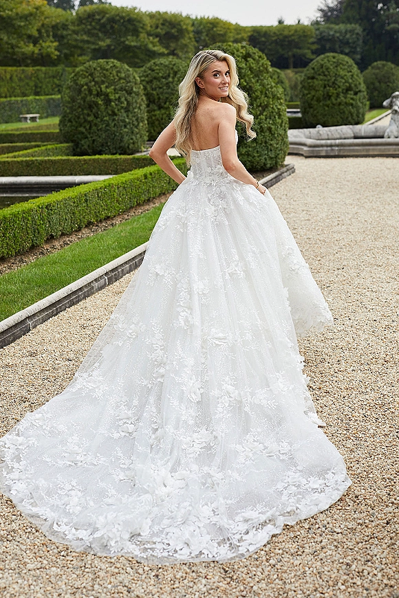 Bridal portrait of a bride in a strapless ball gown with lace appliques and long train, looking over her shoulder by a stone fountain in a formal garden