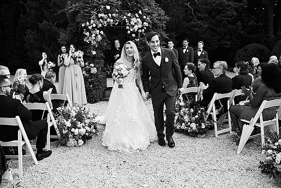 Ceremony recessional as bride and groom walking aisle, kissing beneath a floral arch on a gravel garden path, guests clapping behind