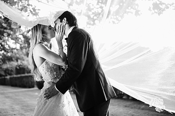 Wedding kiss as bride and groom kissing under a windblown veil, her hand on his face in sunlit greenery outdoors
