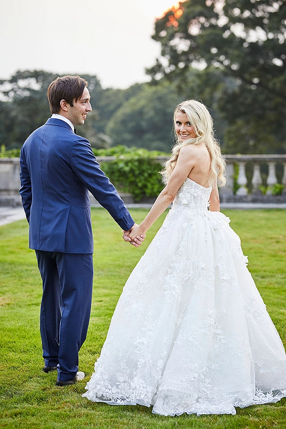 Couple portrait of bride and groom holding hands, walking away on a lawn as she looks back, her lace train beside a stone balustrade