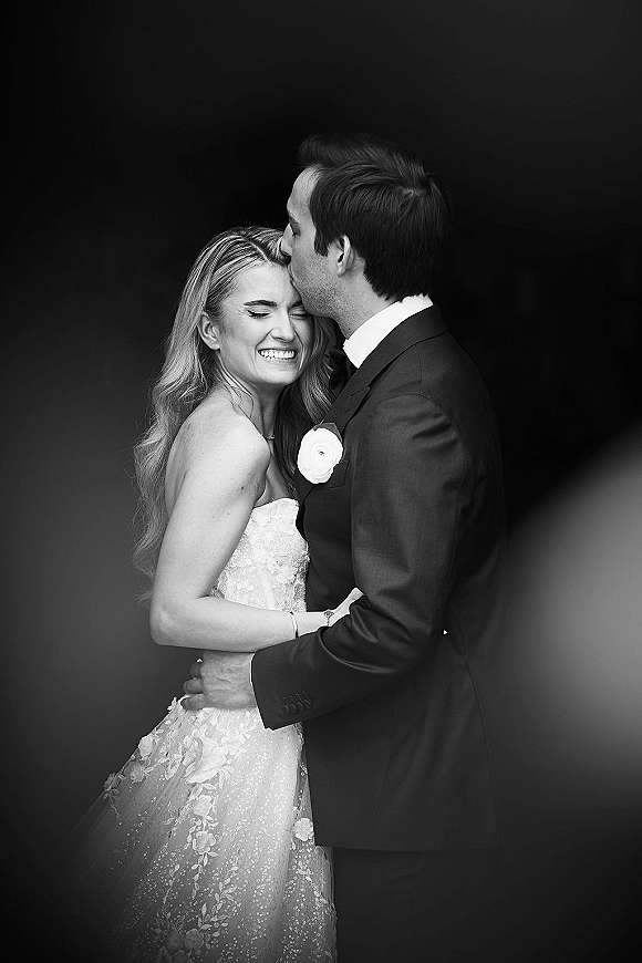 Wedding couple portrait in black and white with groom kissing the bride’s forehead as they embrace against a dark backdrop, lace dress visible