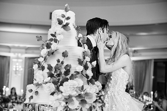 Wedding cake cutting as bride and groom in a strapless gown and tuxedo slice a tiered cake with rose accents in a ballroom reception
