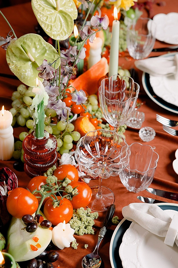 Reception tablescape with family style wedding table featuring taper candles, orchids and anthurium with grapes, citrus, goblets, and plates on wood table